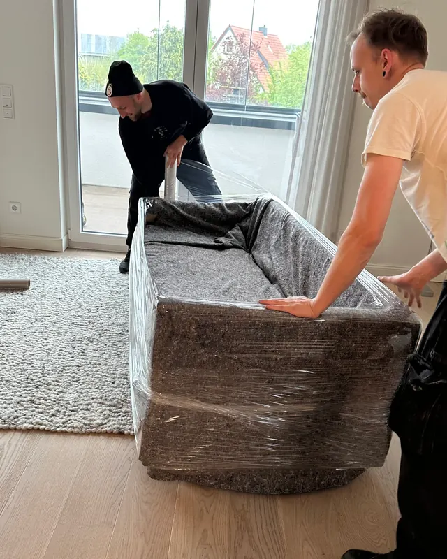 Zwei Männer tragen ein geschütztes Sofa durch einen Wohnungsflur